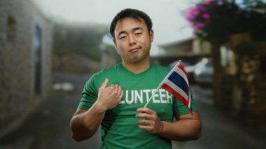 Sokaklarda Tayland bayrağı taşıyan genç adam gönüllü tişört giyiyor. Şehir ortamında kamu hizmeti öneriyor.