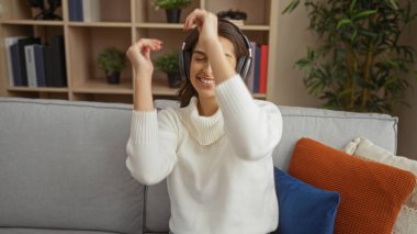 Kadın evde kulaklık takarak neşeyle dans ediyor, rahat bir oturma odasında minderlerle gülümsüyor, modern kapalı alanlarda mutluluk ve rahatlama sergiliyor..