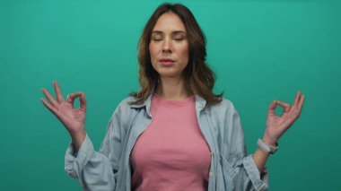 İzole edilmiş yeşil arka plana karşı sakin bir şekilde meditasyon yapan bir kadın. Gözleri kapalı, günlük kıyafetlerini giymiş, sakin bir şekilde farkındalık egzersizi yapan bir kadın.
