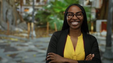 Şehir ortamında gülümseyen, gözlük takan, sarı gömlekli kıvırcık saçlı, kendine güveni ve mutluluğu şehrin arka planında gösteren bir kadın..