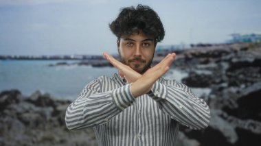 Sakallı İspanyol bir adam kumsalda kollarını çapraz yapıp deniz kenarında bir jest yapıyor..