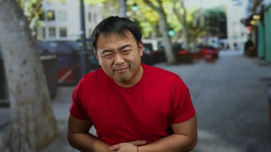 Şehirdeki sokak ortamında midesini tutan kırmızı gömlekli bir adam bulanık bir şehir geçmişine karşı rahatsızlığını dile getiriyor..