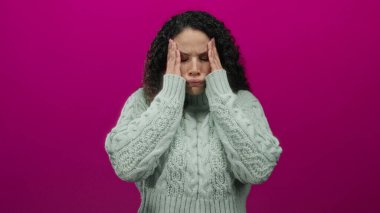 Stresli hisseden kadın, gerilim ve konsantrasyon ifadesiyle çelişen canlı pembe bir arka plana karşı kazak giyerek başını tutuyor..