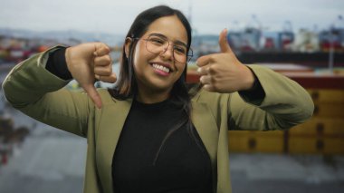 İskele arka planında baş parmak hareketi yapan, gülümseyen, gözlüklü, yeşil ceketli, açık havada kararsızlık ve karışık duyguları yakalayan bir kadın..