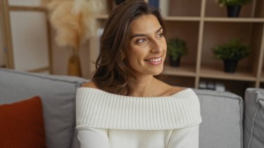 Esmer kadın rahat oturma odasında modern dekor ile gülümsüyor, konforlu kanepe ve bitkilerle dolu raflar, gündüzleri sıcaklık ve rahatlama iletiyor.