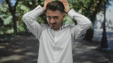 Kulak damızlığı olan beyaz gömlekli adam, sokakta yeşil ağaçların arasında ellerini kaldırır..