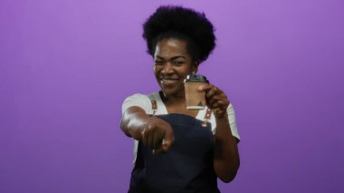 Önlüklü, gülümseyen ve elinde kahve fincanı tutan bir kadın stüdyo ortamında canlı mor bir arkaplan, pozitiflik ve nişan sergiliyor..