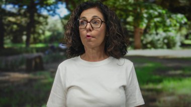 Orta yaşlı İspanyol bir kadın yeşil bir açık hava parkında gülümsüyor, beyaz bir gömlek giyiyor, güneş ışığı ve ağaçlarla çevrili, canlı ve neşeli bir atmosfer yaratıyor..