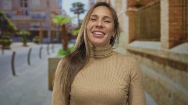 Şehir sokaklarında gülümseyen genç bir kadın. Üzerinde kahverengi bir kazak ve bulanık bir şehir geçmişi var.