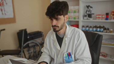 Klinikteki genç adam bilgisayarında tıbbi ekipman ve tekerlekli sandalyeyle çalışıyor. Hastane ortamında sağlık uzmanı olduğunu gösteriyor.