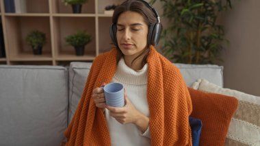 Kadın rahat bir oturma odasında, kulaklıkla ve kahveyle, turuncu battaniyeye sarılmış, modern dekor ve huzurlu atmosferle çevrili, evde sakin vakit geçiriyor..