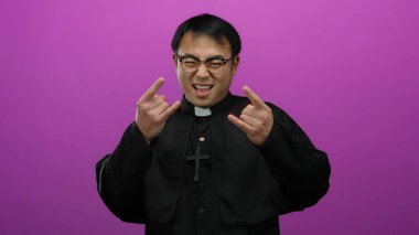 Pembe arka planda neşeli bir şekilde poz veren rahip, taş el işaretini gülümseyerek gösteriyor.