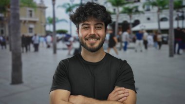Palmiye ağaçları ve binaların yer aldığı kalabalık bir sokakta kollarını kavuşturup güvenle gülümseyen genç İspanyol adam..