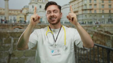Doktor önlüğü ve steteskop giymiş sakallı genç adam, gülümsüyor ve Roma harabelerinin önünde yukarı doğru işaret ediyor..