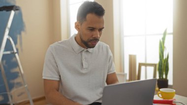Genç İspanyol adam dizüstü bilgisayarda çalışıyor. Yeni bir ev dekorasyonu ve oturma odasında yeni boyalar var..