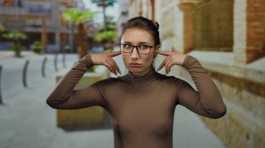 Hayal kırıklığına uğramış bir ifadeyle gözlük takan genç bir kadın şehir sokaklarında kulaklarını tıkıyor.