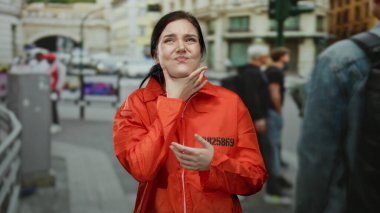 Turuncu hapishane tulumu giymiş bir kadın, rahat hissetmiyor, arka planda insanlarla dolu bir caddede duruyor, açık hava manzarası sergiliyor..