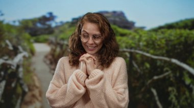 Parkta gülümseyen genç bir kadın rahat bir kazak giyiyor, canlı yeşillik ve doğal yollarla çevrili..