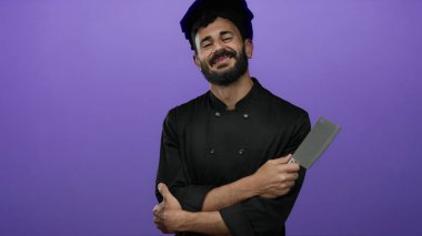 Sakallı bir şef mor arka planda duruyor. Kollarını kavuşturmuş, kendine güveni ve profesyonelliği parlak siyah üniformasıyla yayıyor..