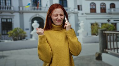 Binanın dışında telefonla konuşan, sarı kazak giyen, şehirde duygularını ifade eden, iletişim yaşam tarzını ve şehir deneyimini sembolize eden bir kadın.