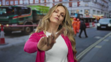 Kendine güveni tam bir kadın inşaat makinelerinin yanında sessiz reddetmeyi işaret ederken sarışın genç bir gözlemci şehir merkezindeki bir cadde boyunca dışarıda duruyor..