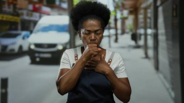 Kalabalık şehir sokağında arka planda araçları olan önlük giymiş bir kadın öksürüyordu. Bu da iş stresi ve kirliliği ön plana çıkaran bir şehir ortamını işaret ediyordu..