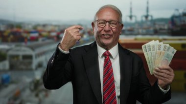 Takım elbiseli kıdemli bir adam, bir sahil limanında Czech Koruna banknotları tutuyor, bir gülümseme ile işaret ediyor, bir denizcilik arka planına karşı başarı ve zenginliğin özünü yakalıyor..