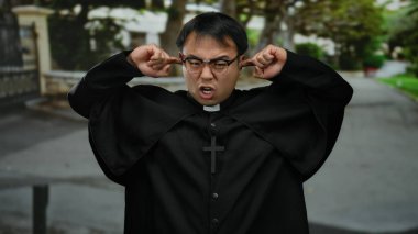 Gözlüklü ve büro kıyafeti giyen bir adam sessiz bir sokakta dikiliyor, elleri kulaklarını haç kolyesiyle kapatıyor, sıkıntı ve hüsranı iletiyor..