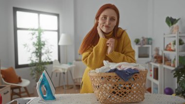 Kızıl saçlı kadın çamaşırlarını çamaşır odasında hasır sepet ve demirle temizliyor.