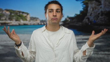 Manzaralı bir sahil sahilinde duygularını ifade eden genç bir adam, açık mavi gökyüzünün altında neşeli bir ruh hali sergiliyor, açık havada beyaz bir gömlekle neşe ve özgürlüğü vurguluyor..