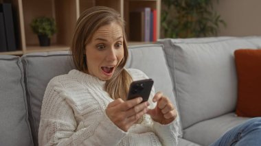Kanepede oturan bir kadın rahat oturma odasında heyecanla telefona bakıyor sıcak ev atmosferini ve kişisel nişanı gösteriyor..