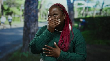 Pembe örgülü, yeşil süveterli, ağaçlarla çevrili bir parkta gülen ve güneşli bir açık hava ortamında doğa ve rahatlığın tadını çıkaran bir kadın..