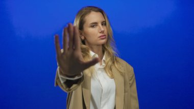 Kadın jestleri, elleri mavi arka planda durur, bej ceket ve beyaz gömlek giyer, temkinli veya ciddi bir ifadeyle uyarır..