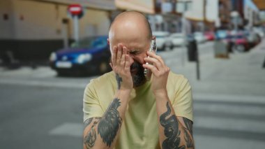 Dövmeli bir adam, şehir caddesinde, kederli bir şekilde telefon tutuyor açık havada konuşurken hayal kırıklığını tasvir ediyor, kel ve sakallı, kederli bir ifadeyle..