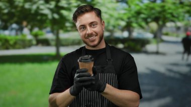 Önlük ve eldiven takan sakallı genç adam güneşli bir açık hava parkında kahve ikram ediyor..
