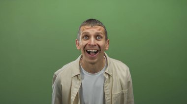 İzole edilmiş yeşil bir arka plan karşısında duran, basit bir bej gömlek içinde çeşitli duygular gösteren, gözleri tamamen açık şaşırmış bir adam..