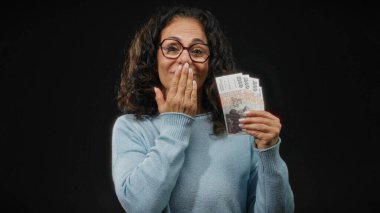 Gözlüklü orta yaşlı bir kadın İzlanda Krona banknotları tutuyor, siyah bir duvara yaslanmış, neşeli bir ifadeyle öpücük yolluyor..