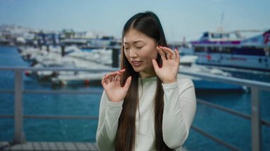Arka planda teknelerle deniz kenarında duran bir kadın, çevreden rahatsız görünüyor, düşünce sahnesini yansıtıyor ve dışarıda huzursuzluk yaratıyor..