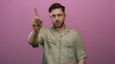 Sakallı genç adam, izole edilmiş pembe bir arka plana karşı onaylanmamış bir jest yapıp bej gömlekli, iddialı bir ifade sergiliyor..