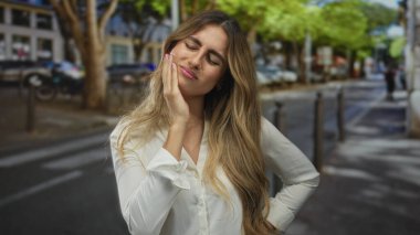 Beyaz bluzlu genç sarışın kadın kaldırımda eliyle yanağına bastırıyor; diş ağrısı, rahatsızlık veriyor..