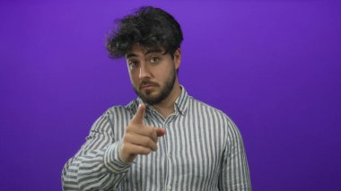 Sakallı genç İspanyol bir adam öne doğru işaret ediyor ve mor arka plan önünde çağrı yapıyor, çizgili gömlek giyiyor, iletişim ve nişan taşıyor..