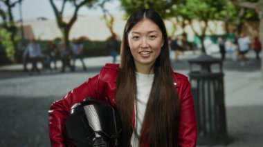 Kırmızı ceketli uzun saçlı kadın, güneşli bir günde ağaçlarla ve arka planda insanlarla şehir sokağında dikilirken miğfer tutuyor..