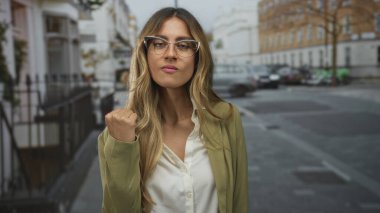 Zeytin rengi ceketli iş kadını, şehir sokaklarında çenesini kaldırıyor; hırs güveni azmi.