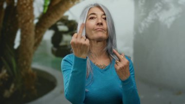 Gri saçlı bir kadın, açık hava ortamında meydan okuma ve yüz ifadesini gösteren saldırgan bir jest yapıyor.