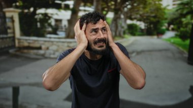 Sakallı İspanyol bir adam, bulanık ağaçlar ve binalarla çevrili bir sokakta sıkıntılı görünüyor, elleri endişeyle ya da hayal kırıklığıyla dolu, güçlü duygular gösteriyor..