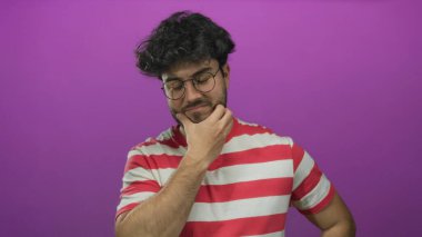 Sakallı ve gözlüklü genç adam pembe arka planda duran çizgili gömlekli düşünceli bir şekilde tefekkür ve içgözlem yapıyor..