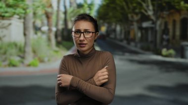 Dışarıda güneş ışığı altında ağaçlarla çevrili bir parkta endişeli gözlüklü bir kadın soğuk sonbahar gününü temsil ediyor..