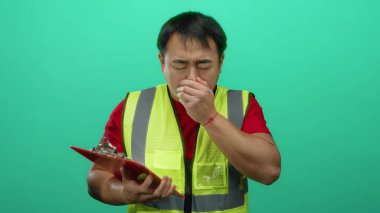 Güvenlik yeleği giymiş genç adam elinde pano tutarak yeşil arka plana hapşırıyor ve işyerindeki sağlık sorununu gösteriyor