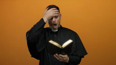 Genç rahip parlak turuncu arka planda sürpriz bir şekilde İncil okuyor, dini bağlılık ve beklenmedik keşifler sergiliyor..