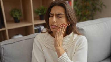 Şık bir oturma odası ortamında diş ağrısı çeken bir kadın yüzüne dokunurken rahatsızlığını belli ediyor. Arka planda iç mekan bitkileri, rahat bir ev ortamını gözler önüne seriyor..